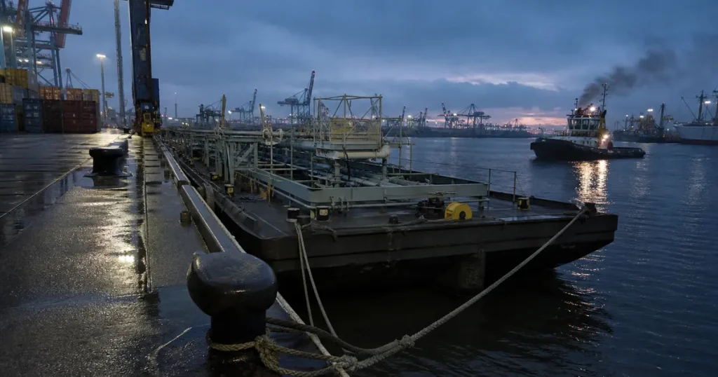 Industrihamn med pråm förtöjd vid kaj i kvällsljus, containerkranar i bakgrunden