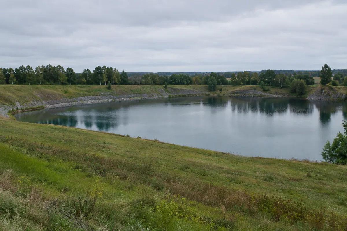 Vattenfyllt före detta dagbrott omgivet av gräsbevuxen täckt mark och lövskog i Västergötland
